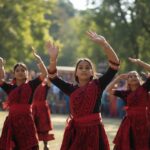 a candid, eye level photograph of a group of young women performing a traditional folk dance outdoors in guwahati. they are wearing red and black woven traditional mising dresses. they have thei (1)