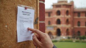 a close up shot of a notice board on a university campus wall. a white paper list titled 'provisional merit list' is pinned to it. a student's hand is pointing at a name on the list. the background
