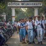 a realistic street shot of students in school uniforms (white and sky blue) walking out of cotton university in guwahati after an exam. some are discussing question papers, others are on their phone